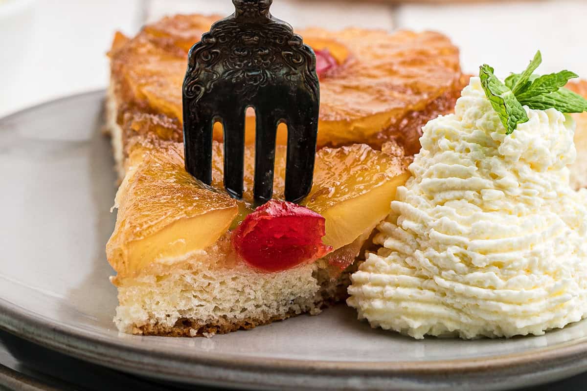 A fork in a slice of pineapple upside-down cake with whipped cream and a mint garnish on the side.