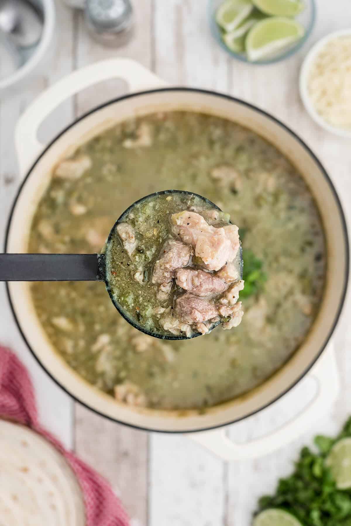 A ladle of green pork chili held over a pot, with limes and cheese in the background.