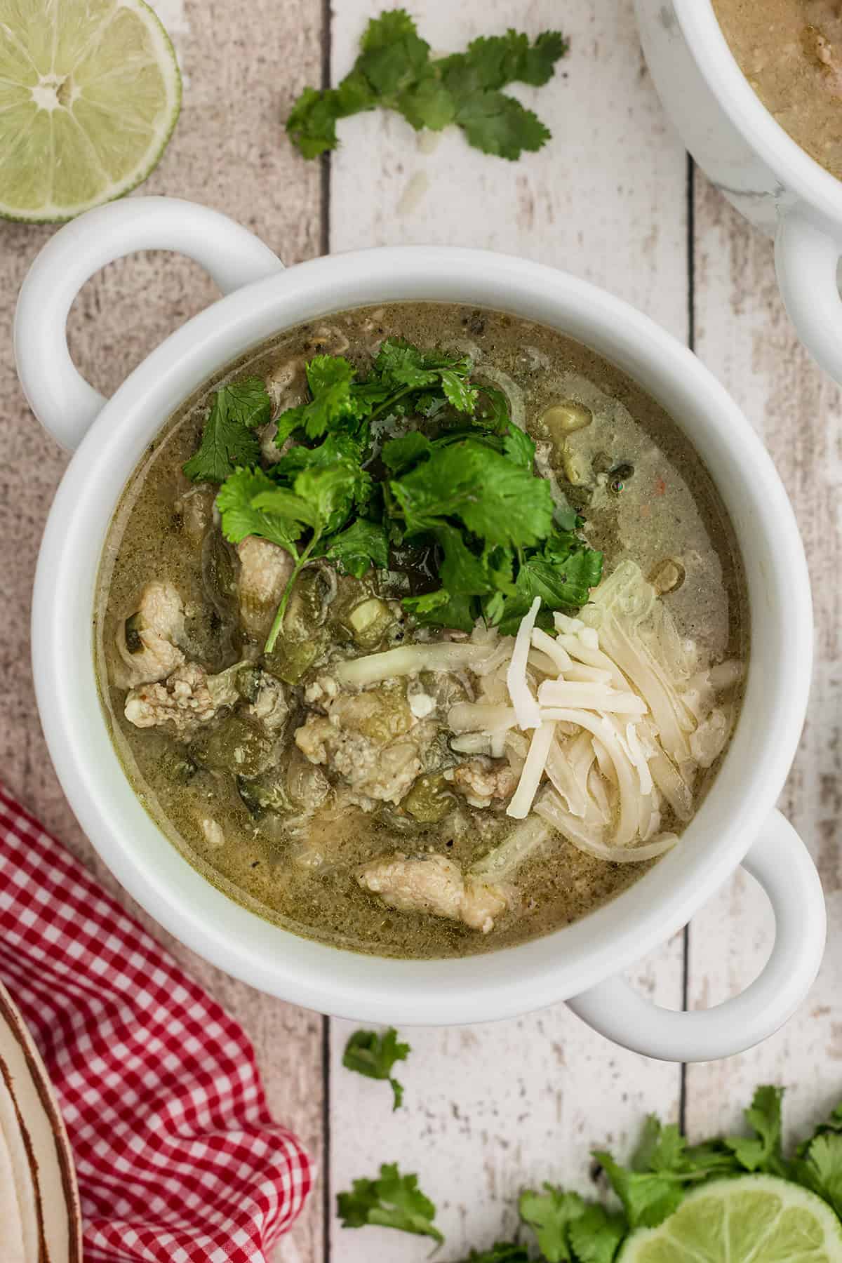 A bowl of green chili stew topped with cilantro and shredded cheese, garnished with lime and a red napkin nearby.