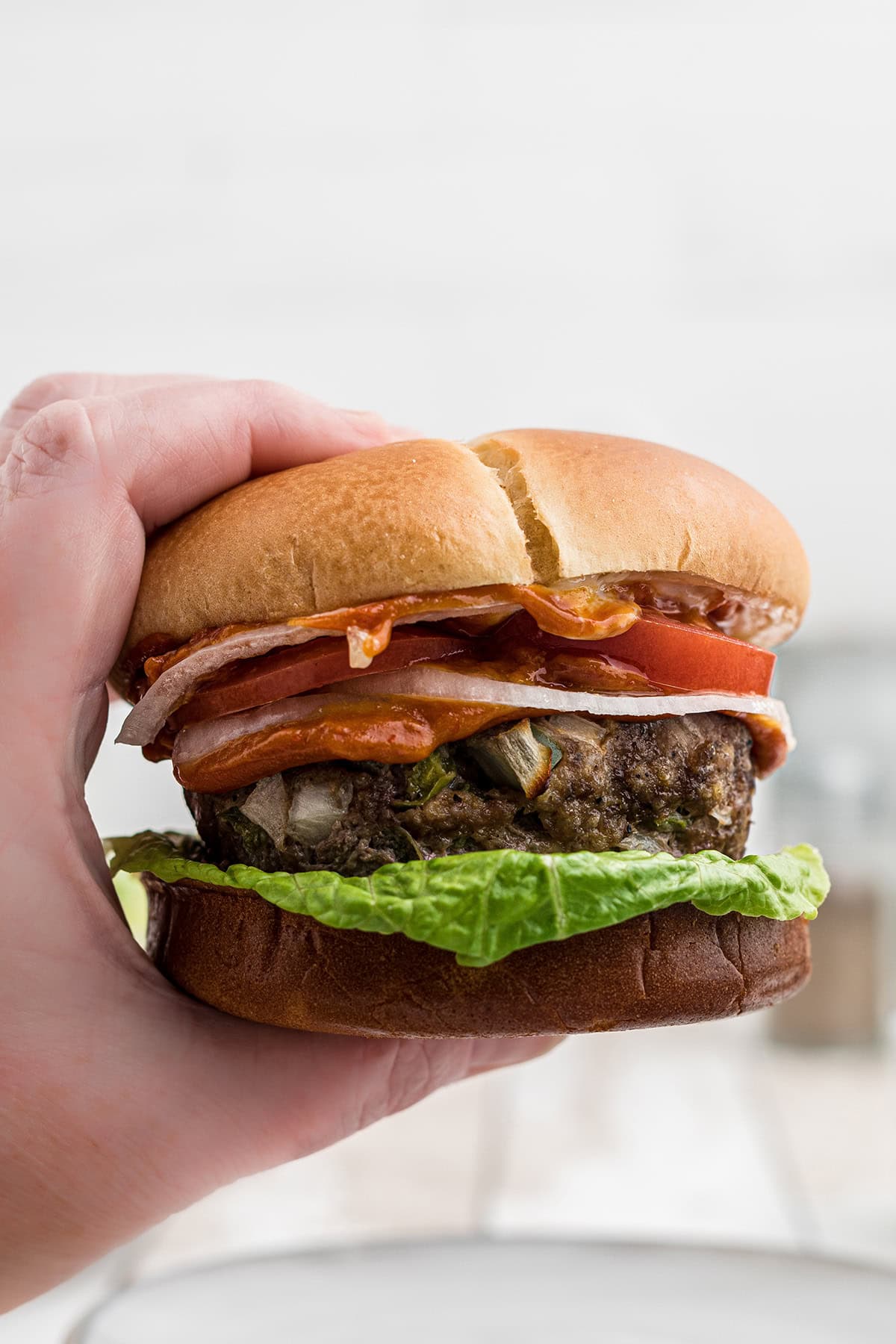 A hand holding a burger with lettuce, onion, tomato, bacon, and a beef patty in a bun.