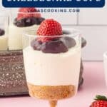 A no-bake cheesecake cup topped with cherry sauce and a strawberry, shown on a pink surface.