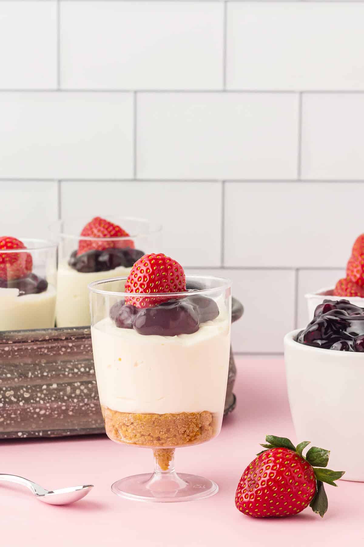 A cheesecake cup with graham crust, cream filling, berry topping, and a fresh strawberry.