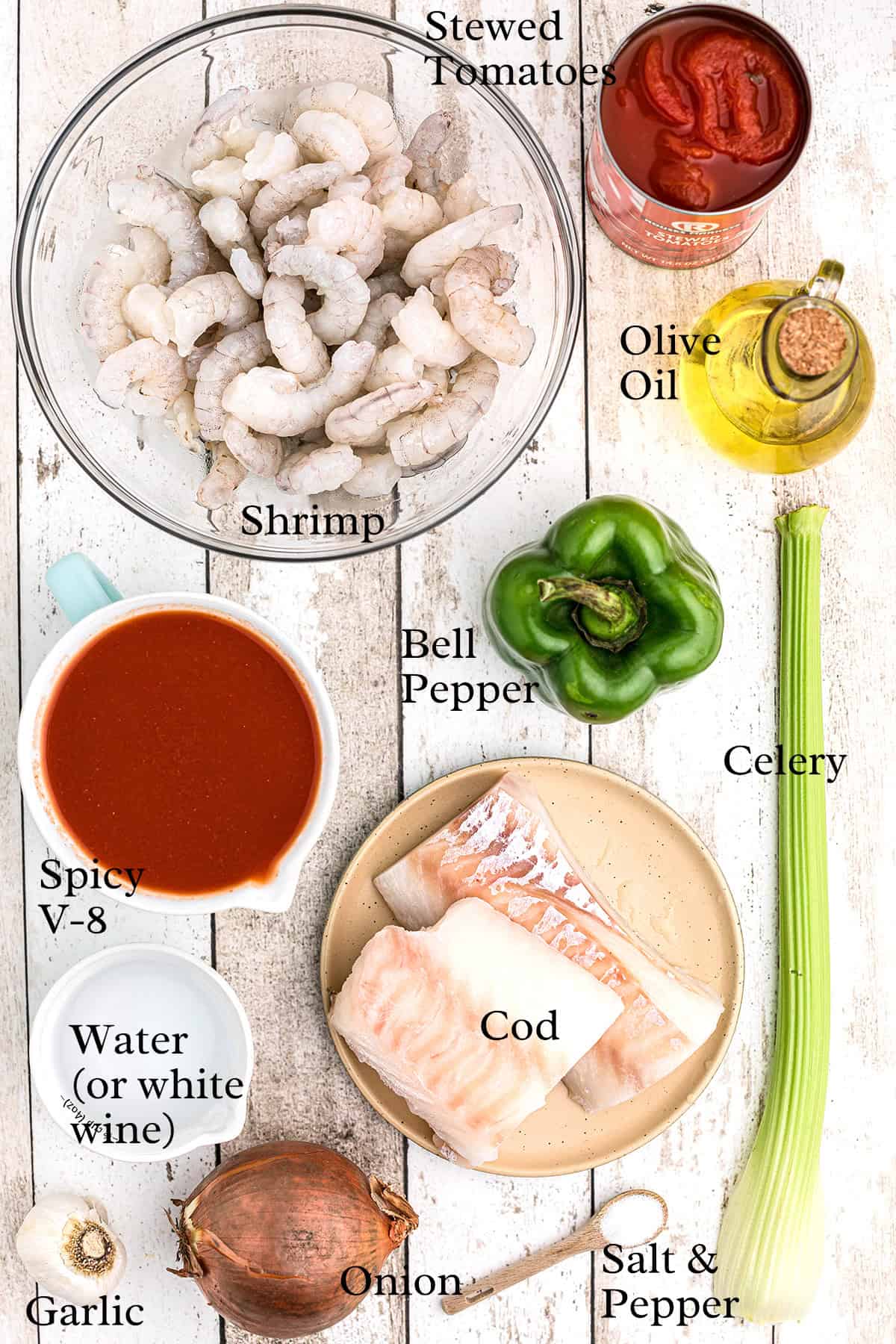 A flat lay of ingredients on a white wooden surface, including raw shrimp, a can of stewed tomatoes, olive oil, a green bell pepper, celery, cod fillets, salt and pepper, an onion, garlic, spicy V-8 juice, and water.