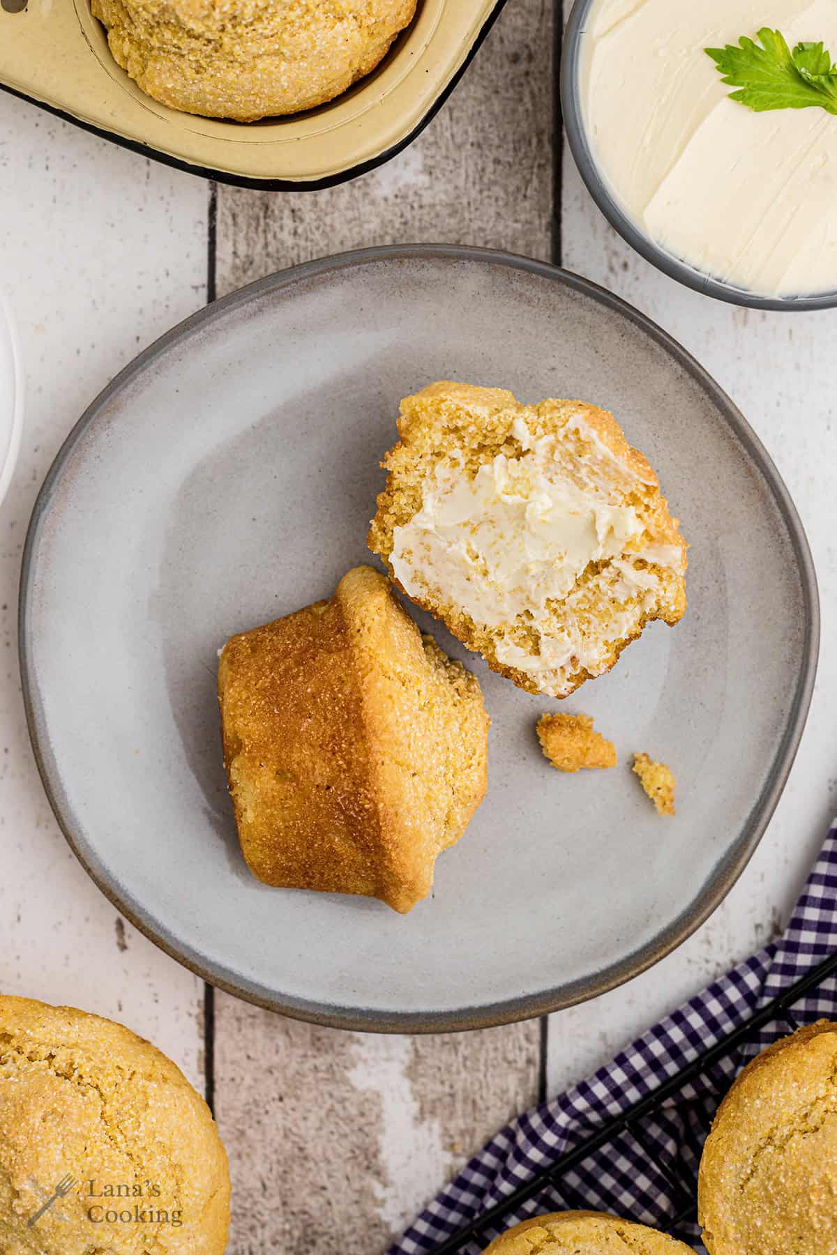 A cornbread muffin split open with butter on a gray plate, beside a whole muffin and a bowl of butter.