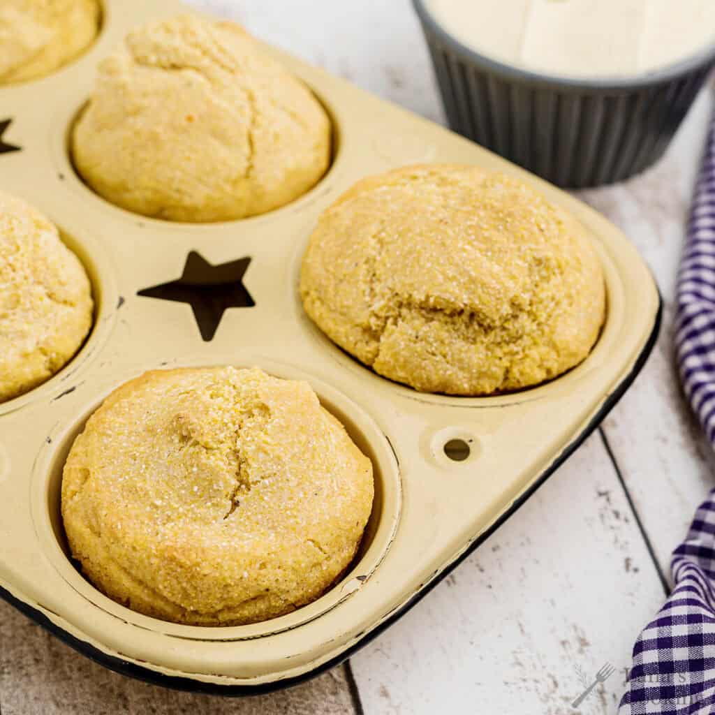 Golden cornbread muffins in a baking tray with butter and a checkered cloth on the side.