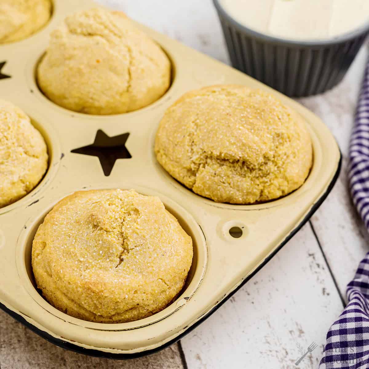 Golden cornbread muffins in a baking tray with butter and a checkered cloth on the side.