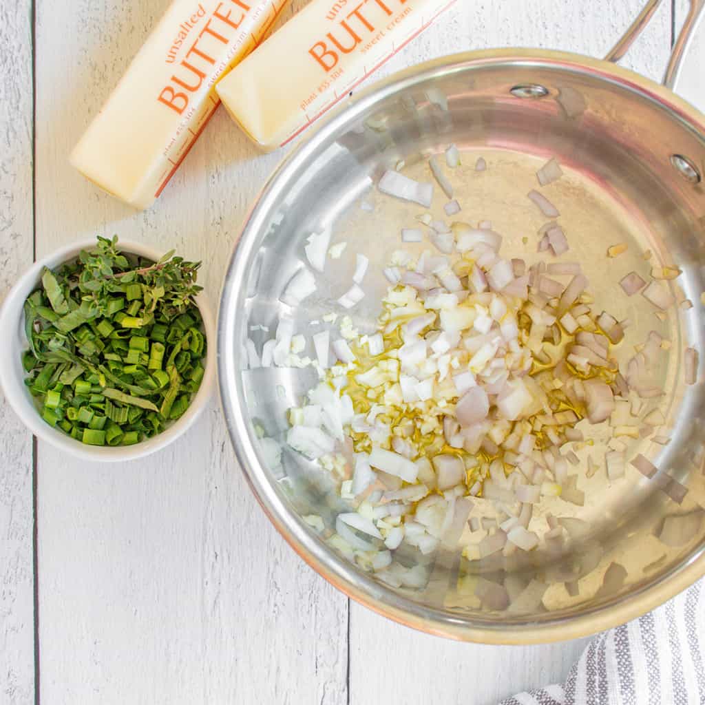 Chopped shallots in a pot, with a bowl of herbs and sticks of butter on a white wooden surface nearby.