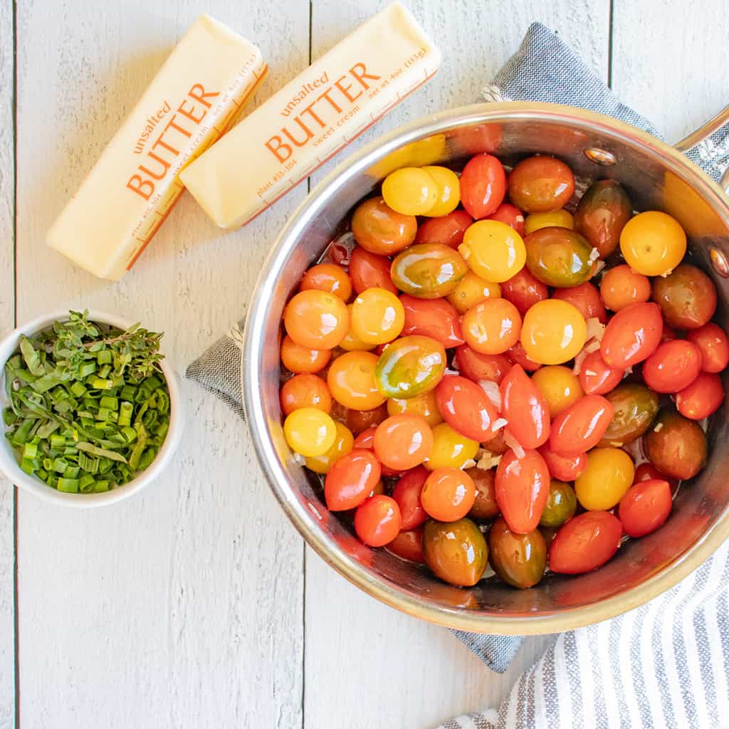 A saucepan of colorful cherry tomatoes next to chopped herbs and two sticks of unsalted butter on a table.