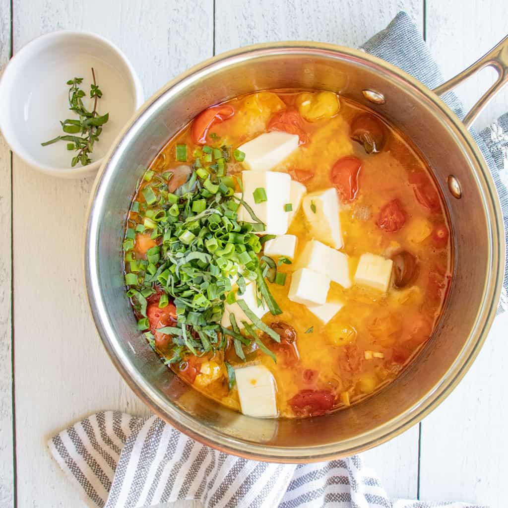 A saucepan of cherry tomato sauce with cubes of butter and chopped herbs, next to a napkin and a bowl of thyme.