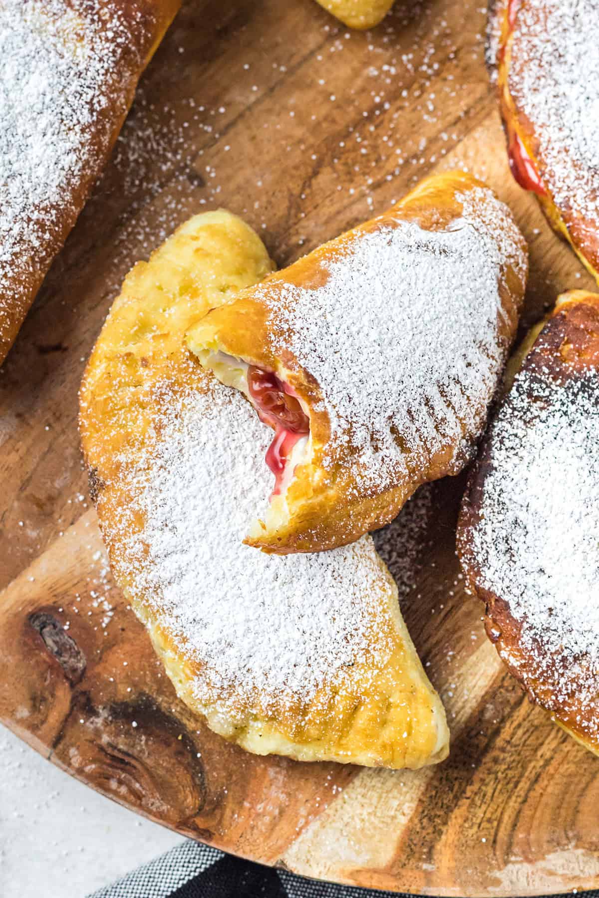 Golden fried hand pies dusted with powdered sugar, one with a bite showing a cherry filling, on a wooden board.