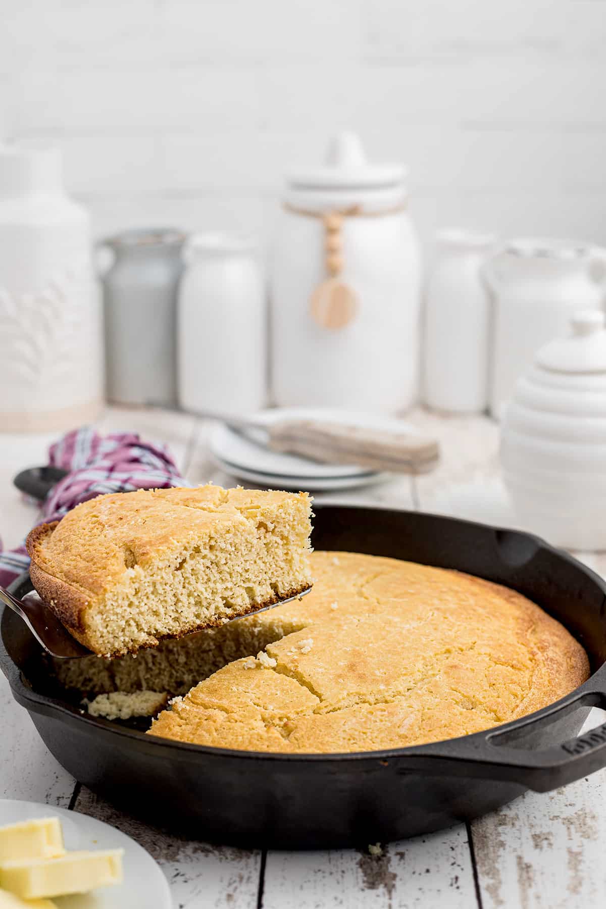 A slice of golden cornbread is being lifted from a cast-iron skillet on a rustic white table.