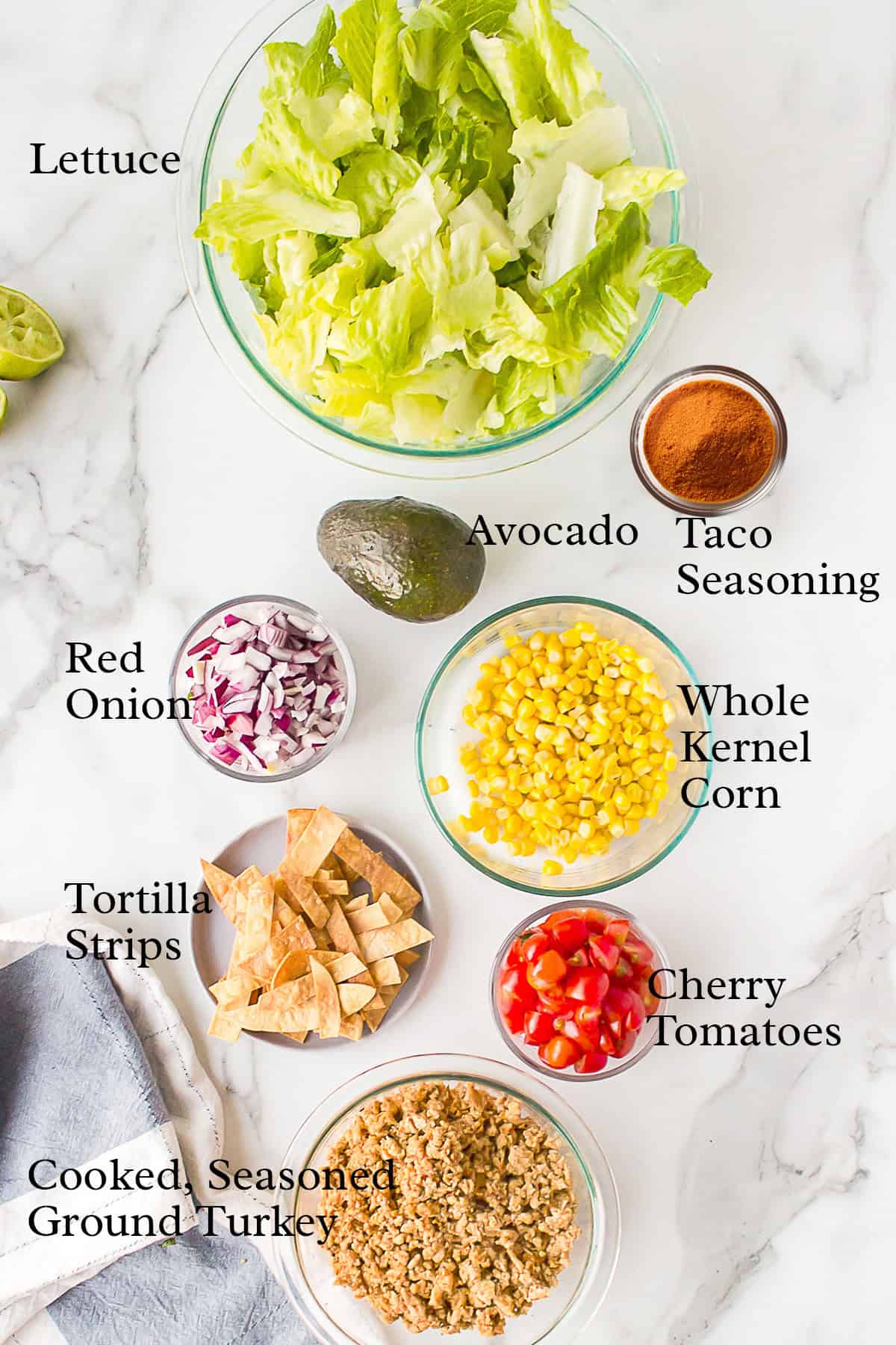 Ingredients for a taco salad arranged in bowls: lettuce, avocado, taco seasoning, onion, corn, tomatoes, turkey, chips.
