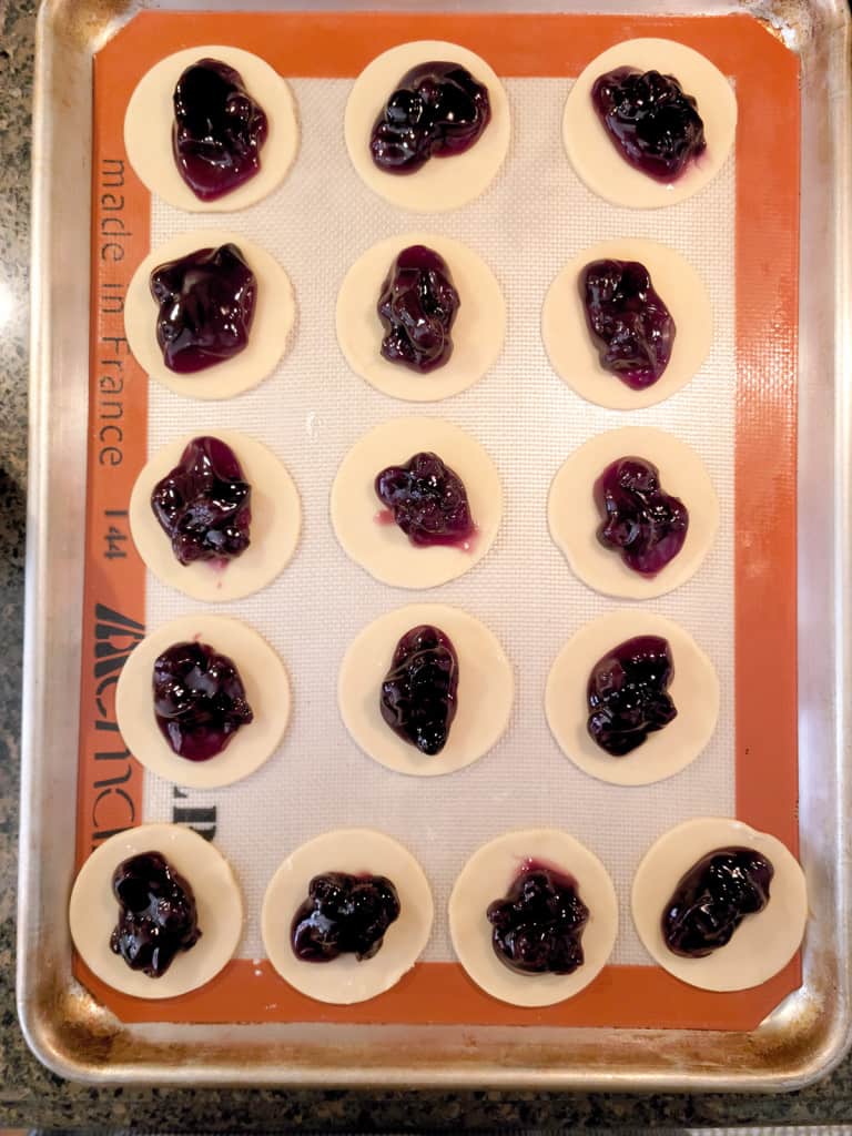 Round dough pieces topped with blueberry filling arranged on a baking sheet lined with a silicone mat.