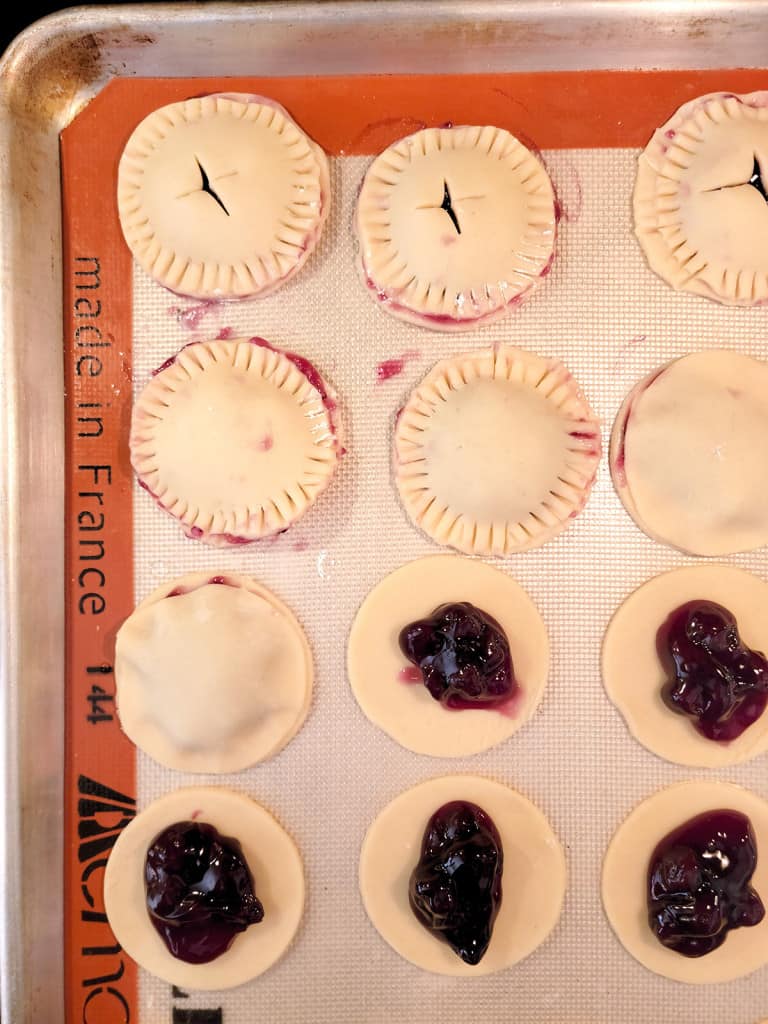 Unbaked round pie cookies with blueberry filling on a baking sheet, some covered and crimped, some open.
