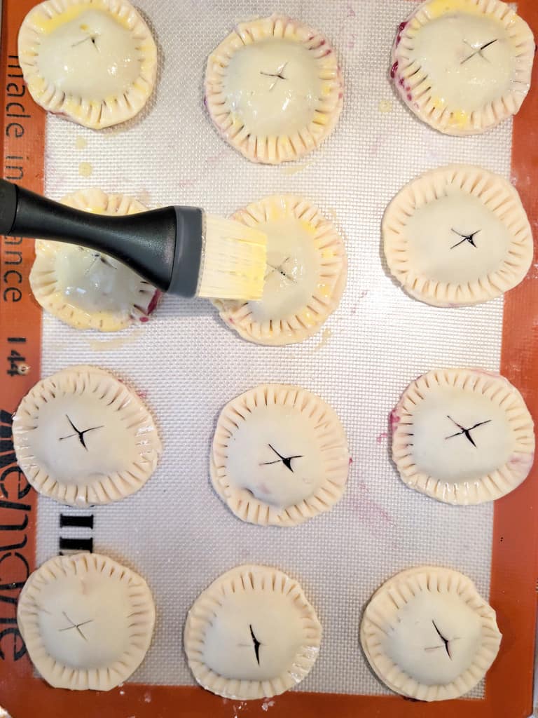 Unbaked blueberry pie cookies on a baking mat, being brushed with egg wash before baking.