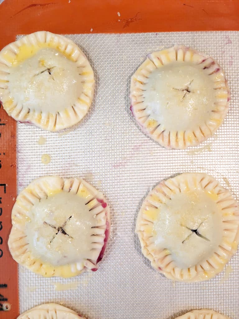 Four unbaked round pie cookies with fork-pressed edges and slits on top, topped with sugar on a baking mat.