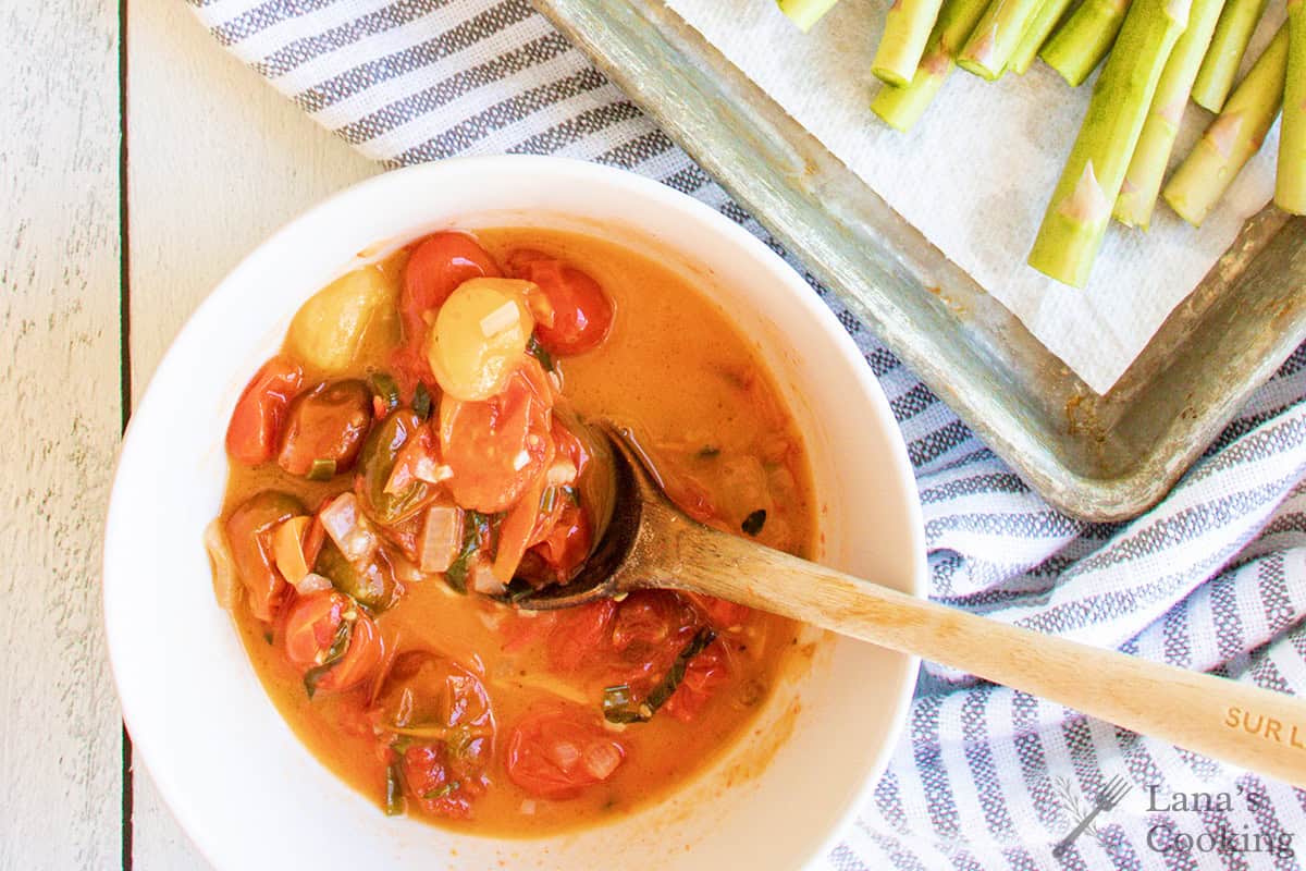 Bowl of tomato-based sauce with vegetables and a wooden spoon, next to a tray of cut asparagus.
