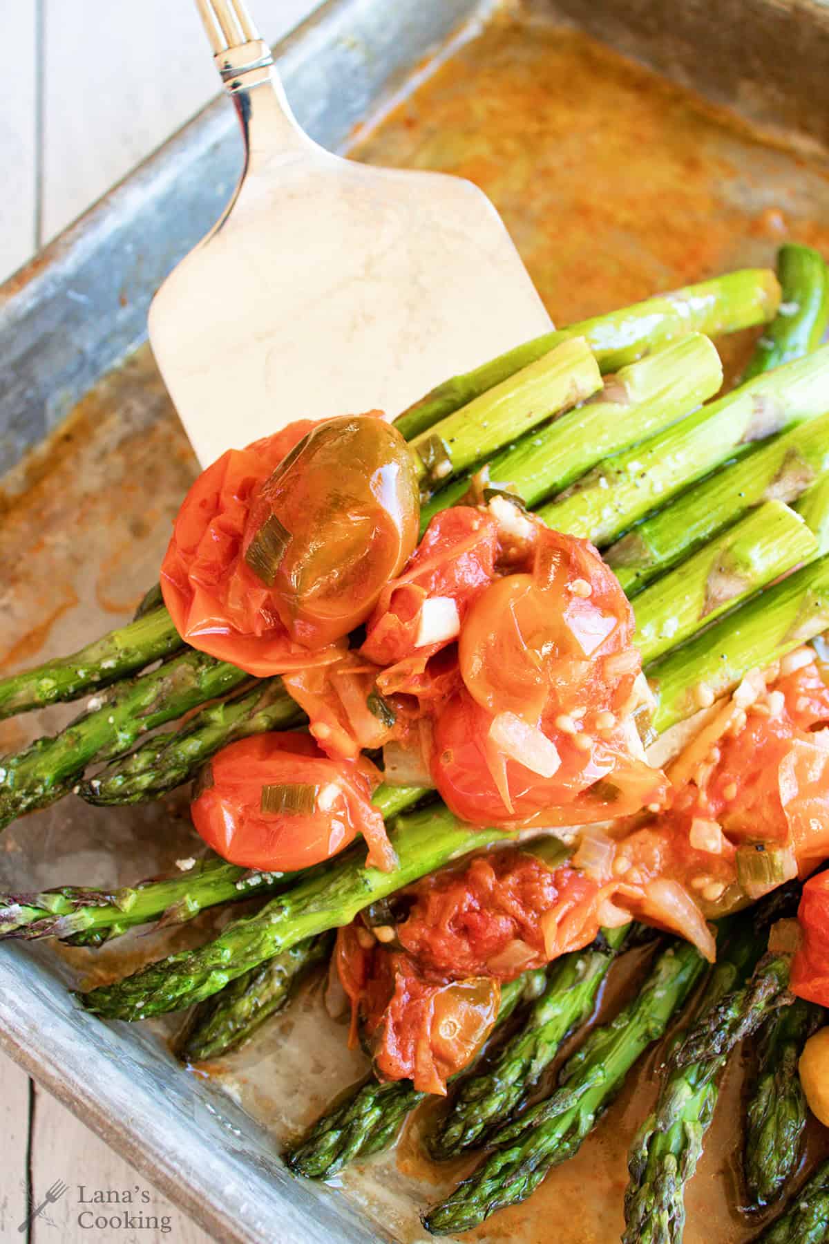 Roasted asparagus topped with cooked cherry tomatoes and onions being served with a spatula.
