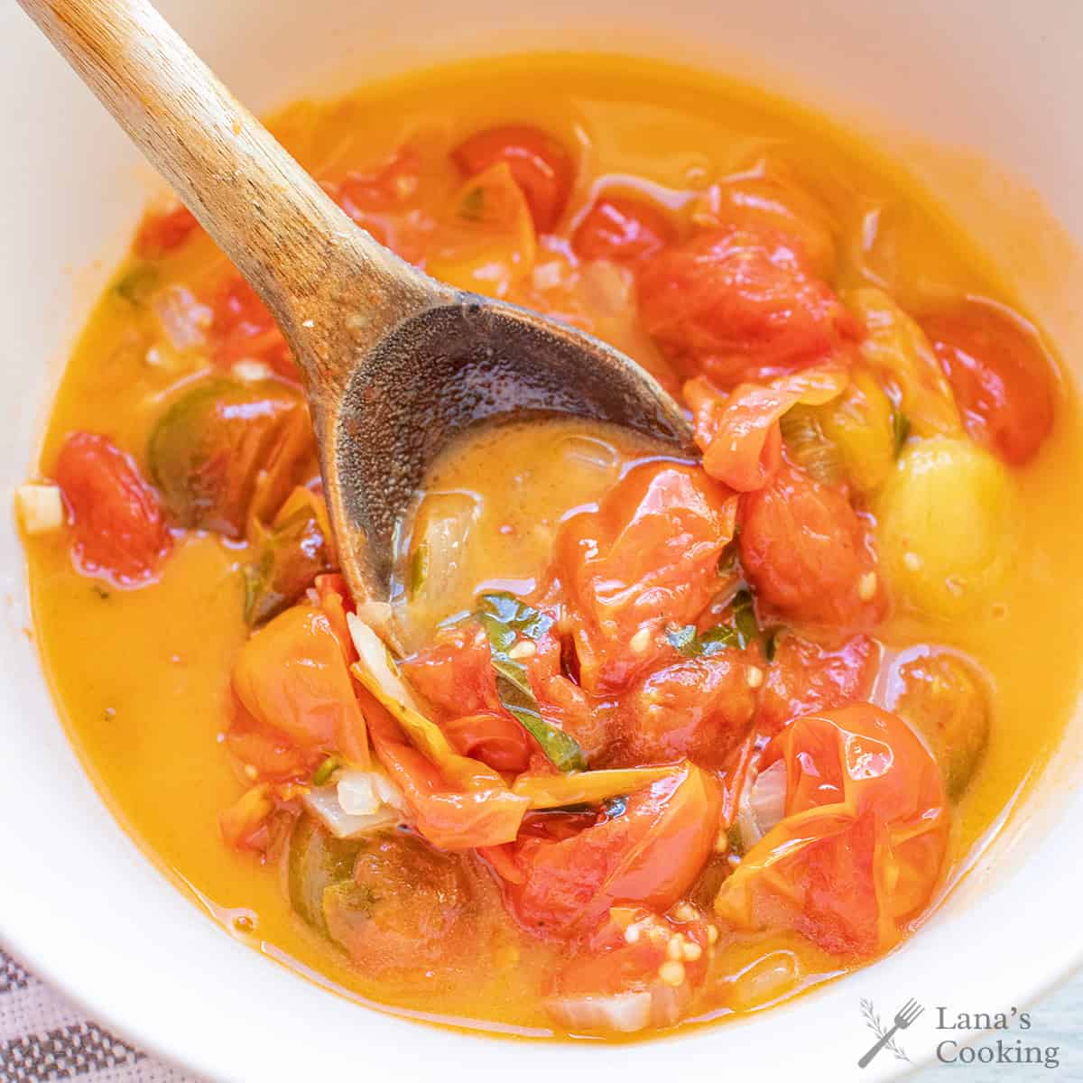 A wooden spoon stirs a bowl of burst cherry tomato sauce in a yellow-orange broth.