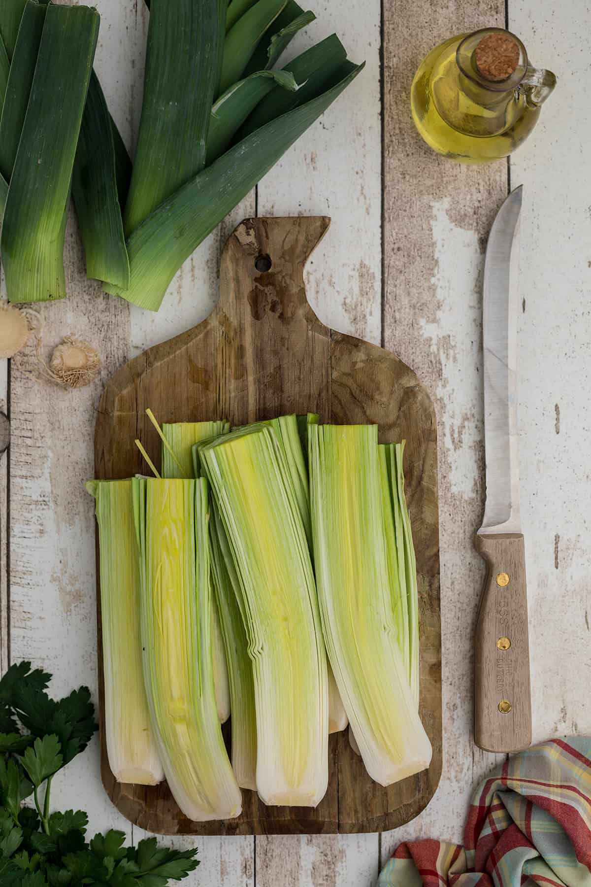 Split leeks on a wooden cutting board with a knife, olive oil, and herbs on a rustic table.