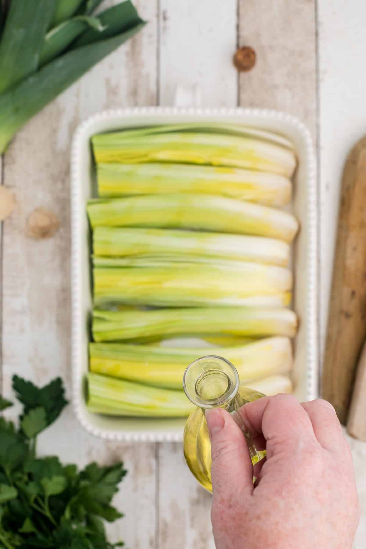 A hand pours oil over halved leeks arranged in a white baking dish on a rustic wooden surface.