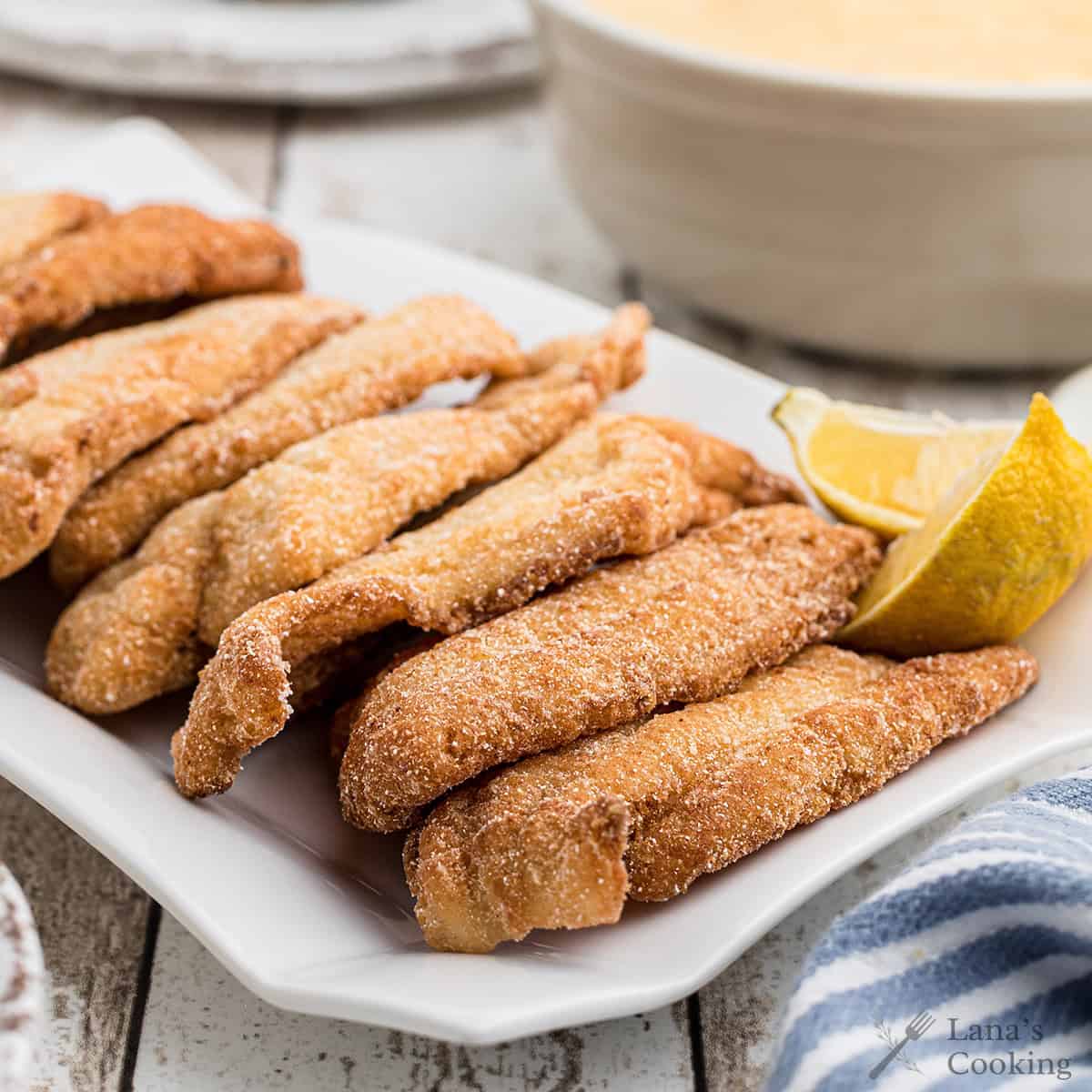 Golden fried catfish fillets on a white plate with a lemon wedge, served on a rustic table.