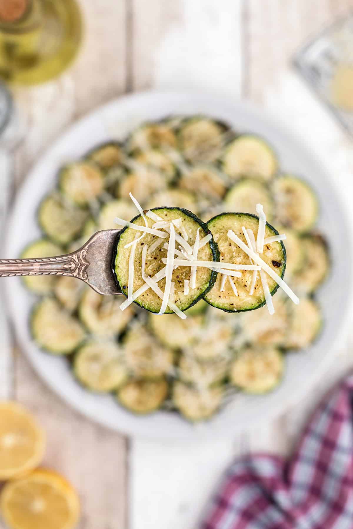 A fork holds two zucchini slices topped with shredded cheese over a plate of zucchini on a rustic table.