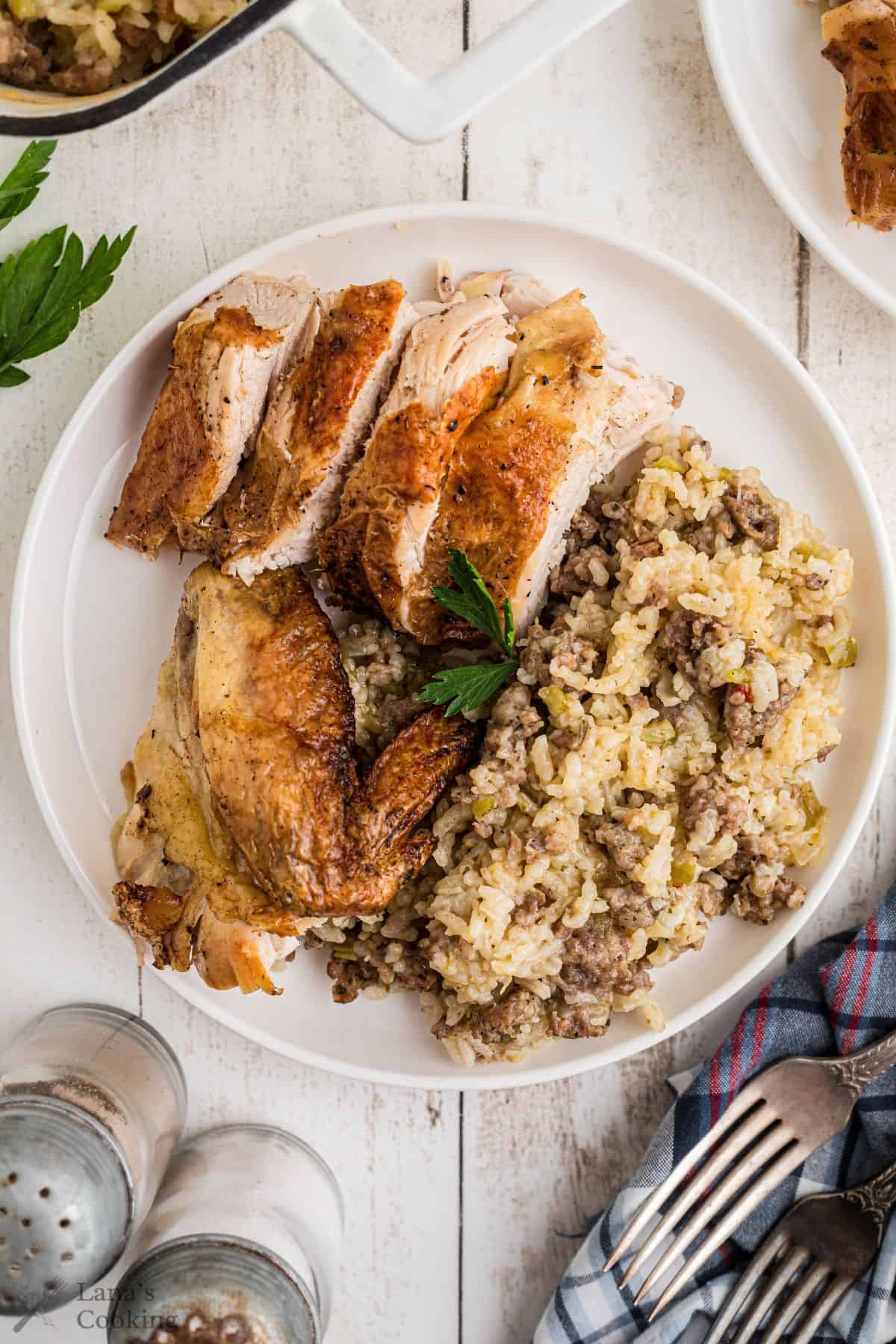 A plate with roasted chicken pieces and a serving of rice mixed with ground meat, garnished with a parsley sprig, on a white table next to salt and pepper shakers.
