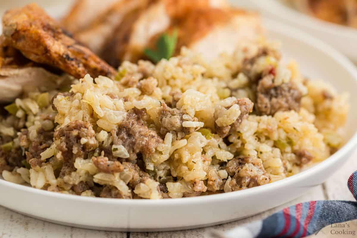 A close-up of a plate of cooked rice mixed with ground sausage and seasonings, with pieces of roasted chicken in the background.