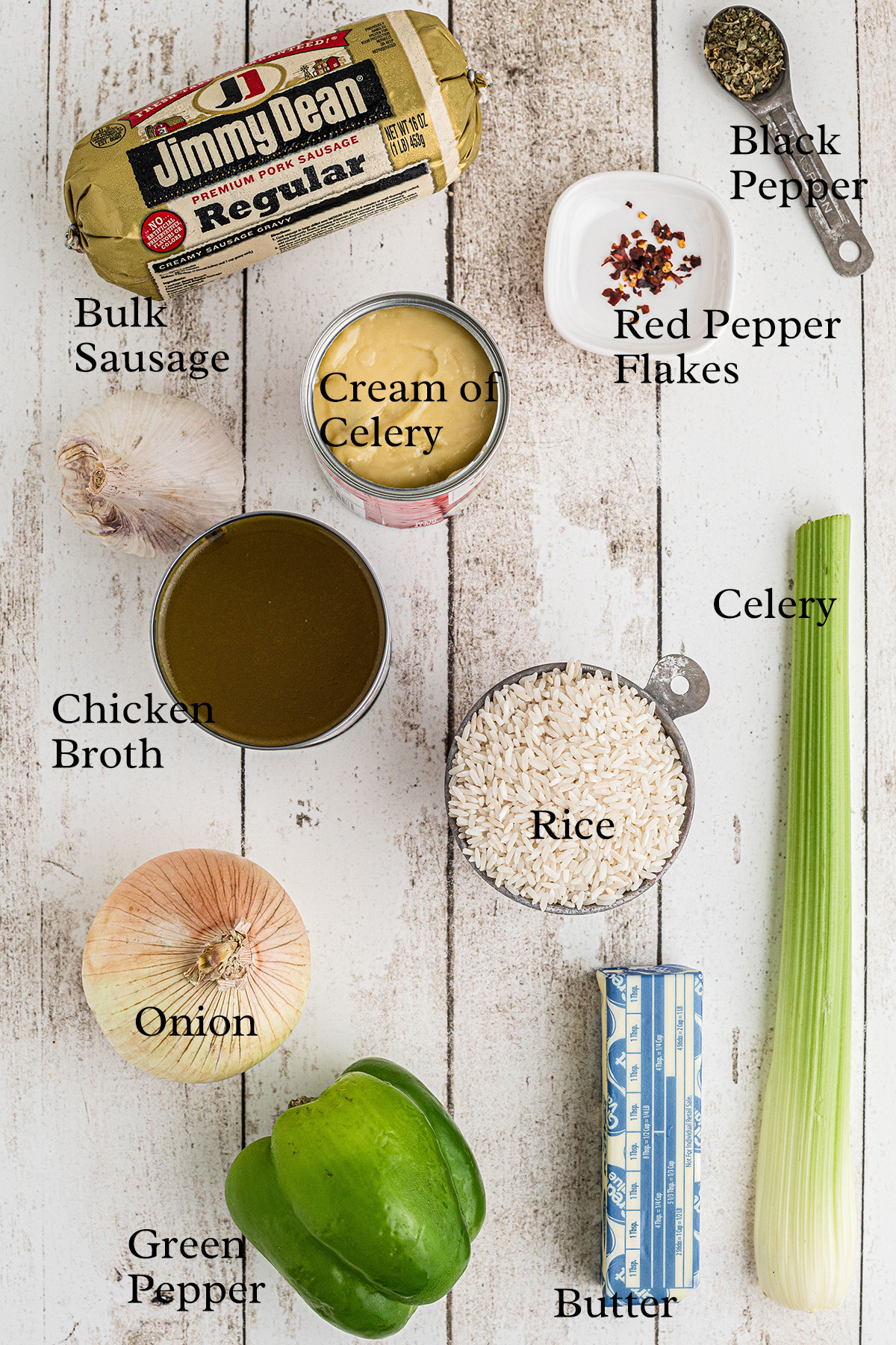 Various ingredients for a recipe are arranged on a white wooden surface, including sausage, cream of celery, spices, broth, rice, celery, onion, green pepper, and butter.