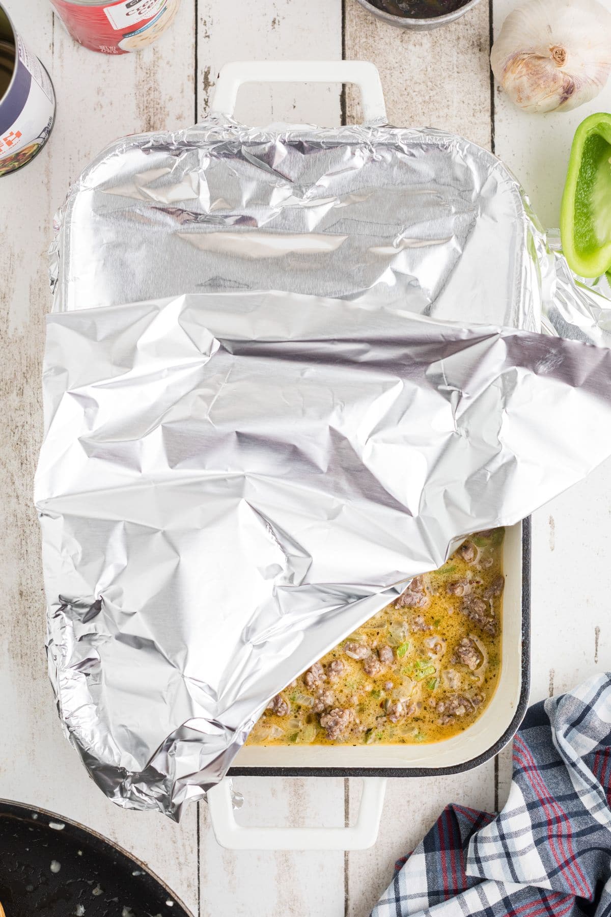 A baking dish partially covered with aluminum foil contains a meat and vegetable mixture; ingredients and kitchen items are visible around it on a white wooden surface.