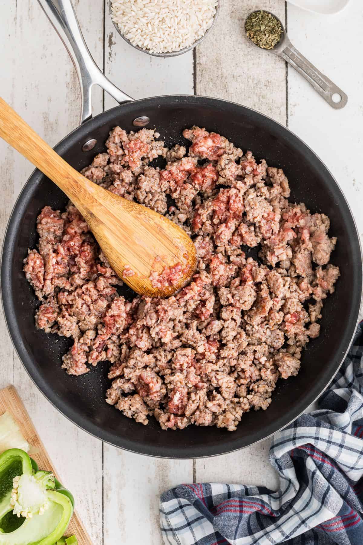 Ground beef is being browned in a black skillet with a wooden spoon on a white wooden surface; raw rice, seasonings, a green bell pepper, and a cloth are nearby.