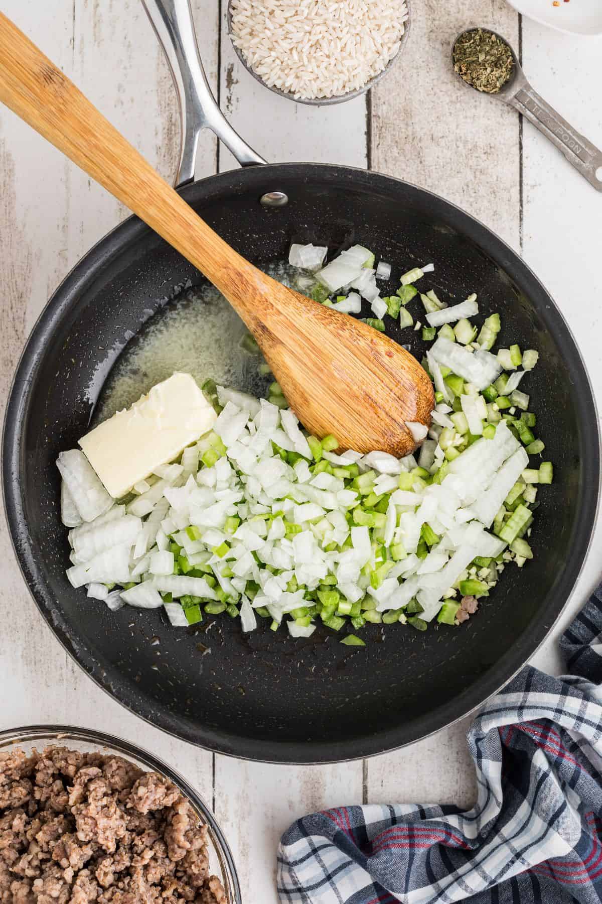 Chopped onions and green bell peppers with a block of butter in a skillet, being stirred with a wooden spoon. Nearby are bowls of ground meat, uncooked rice, and dried herbs.