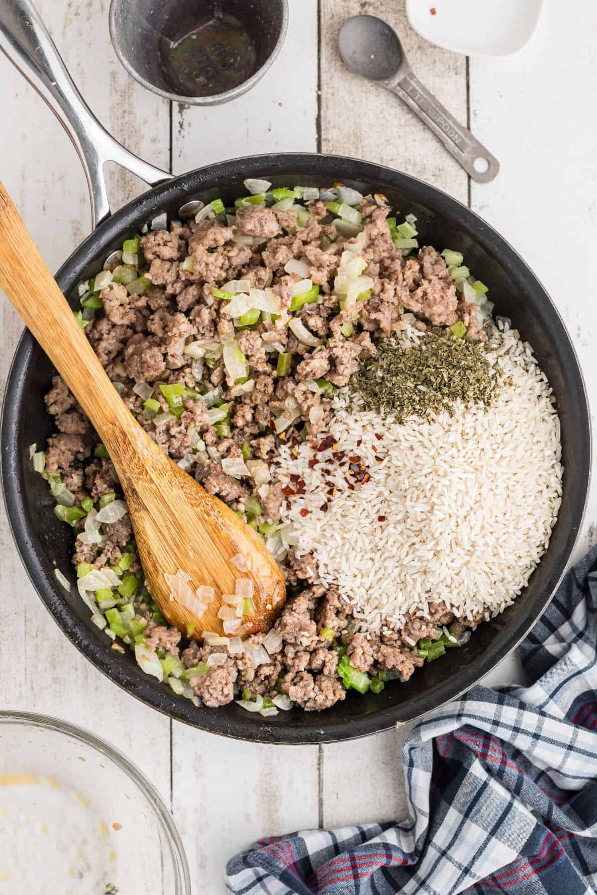 A skillet contains ground meat, chopped onions, celery, herbs, uncooked white rice, and red pepper flakes being mixed with a wooden spoon.