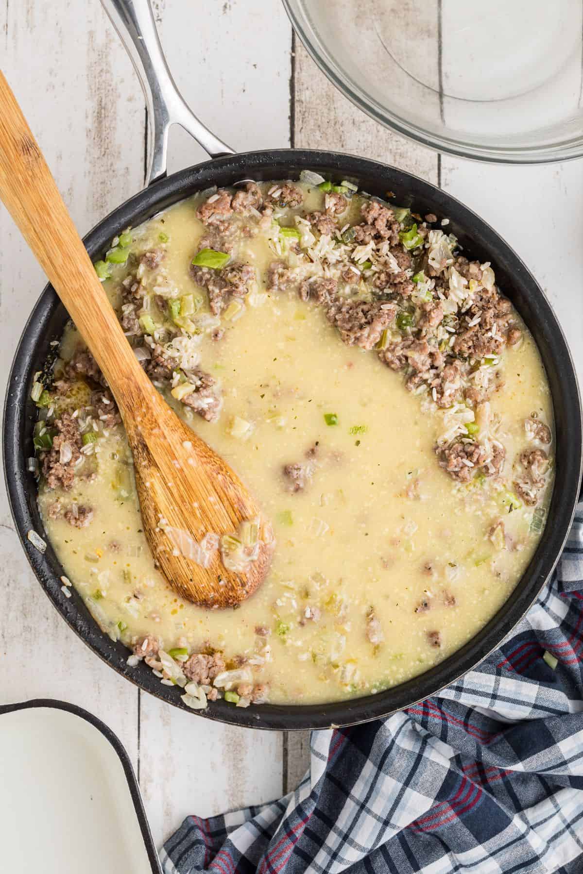 A skillet containing a mixture of ground beef, rice, chopped celery, and broth is being stirred with a wooden spoon. A plaid cloth and empty bowl are nearby on a white wooden surface.