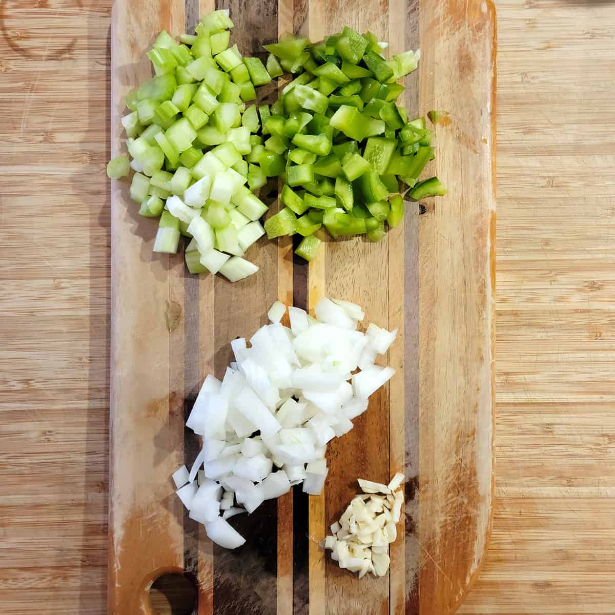 Chopped onion, celery, bell pepper, and garlic on a wooden board.