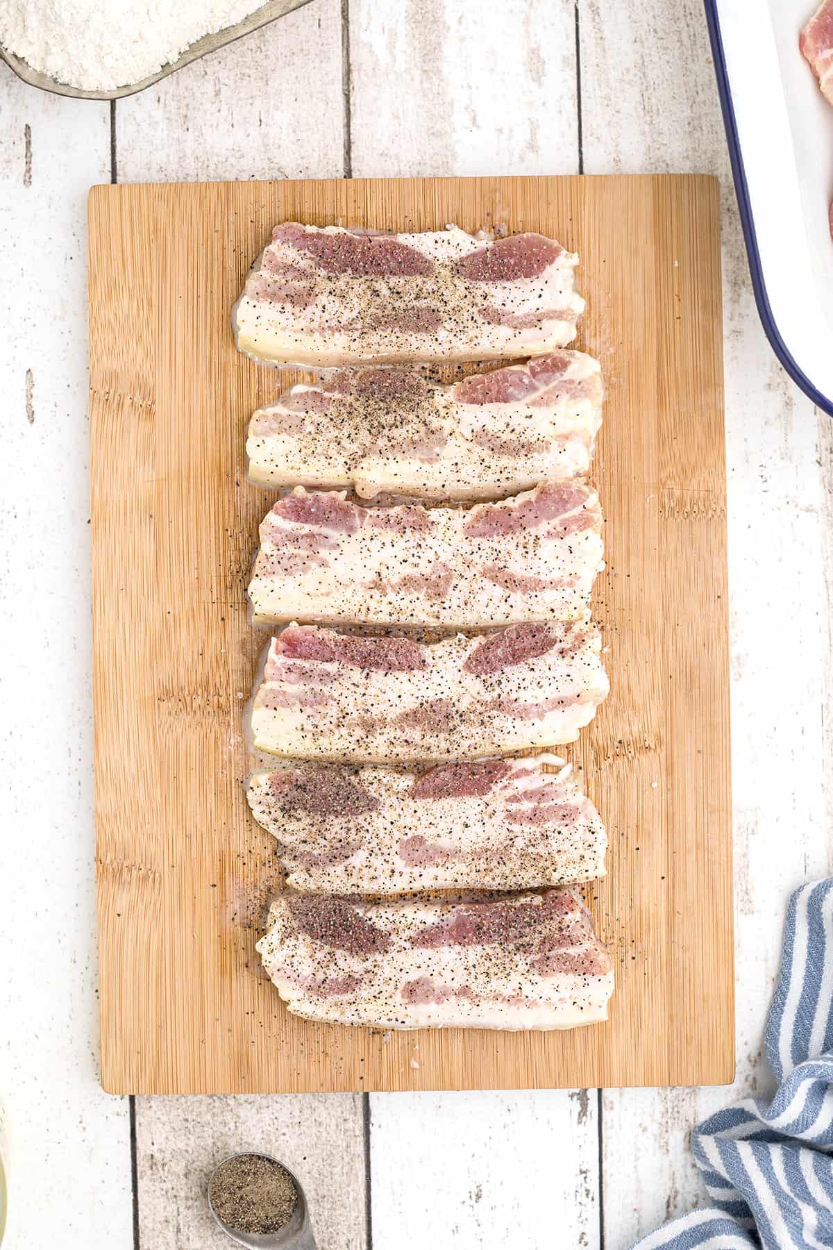 Salt pork slices on a board with ground pepper sprinkled over top.