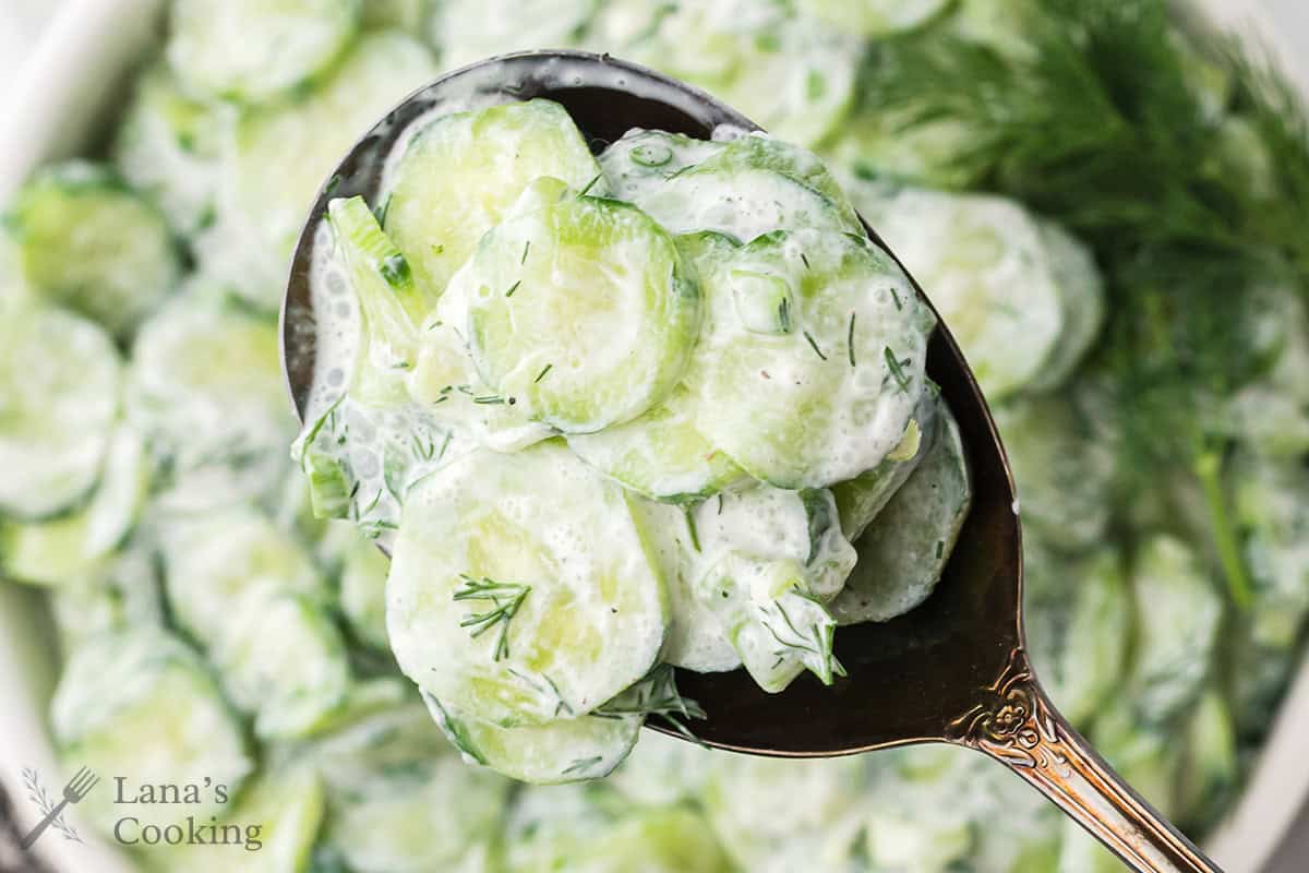 A close-up of a spoon holding creamy cucumber salad with sliced cucumbers and dill. The background shows more of the same salad in a bowl. The image has the text &ldquo;Lana&rsquo;s Cooking&rdquo; in the lower left corner.