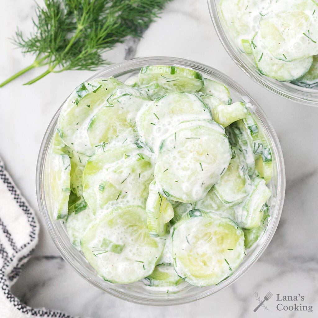 A clear bowl filled with creamy cucumber salad garnished with fresh dill, placed on a marble surface next to a sprig of dill and a black-and-white striped cloth.