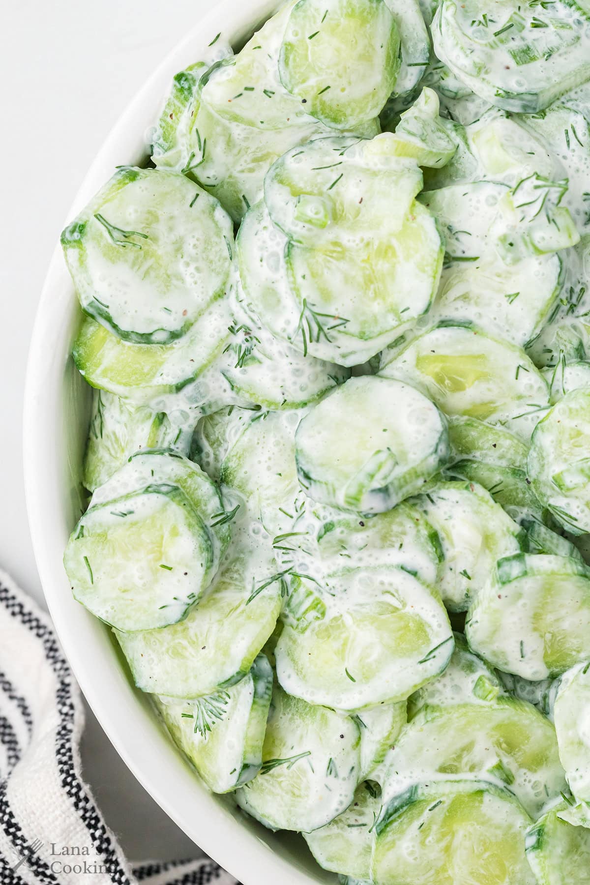 Sliced cucumbers coated in a creamy white dressing with bits of fresh dill, served in a white bowl. A black and white striped cloth is partially visible in the corner.