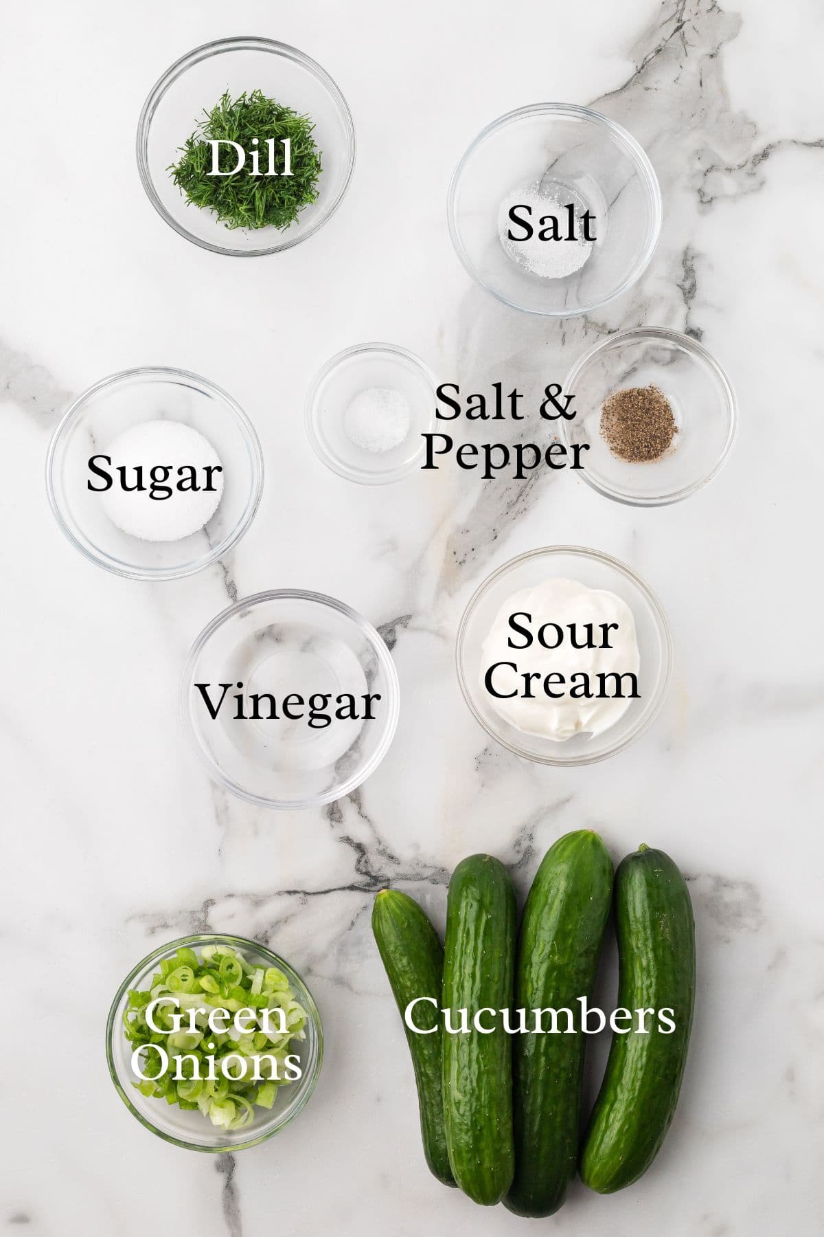 A marble surface with labeled bowls containing dill, salt, sugar, salt & pepper, vinegar, sour cream, green onions, and four whole cucumbers arranged at the bottom right.