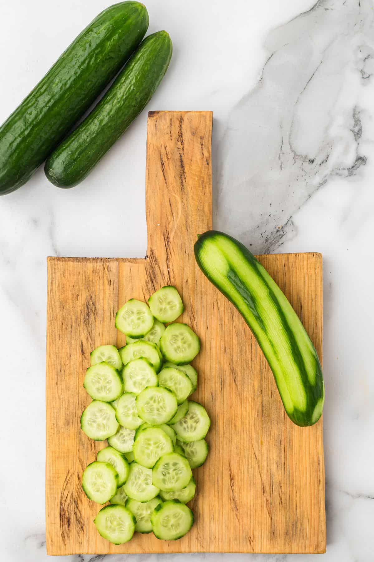 A wooden cutting board on a marble surface holds a partially peeled cucumber and several cucumber slices. Two whole cucumbers are placed beside the board.