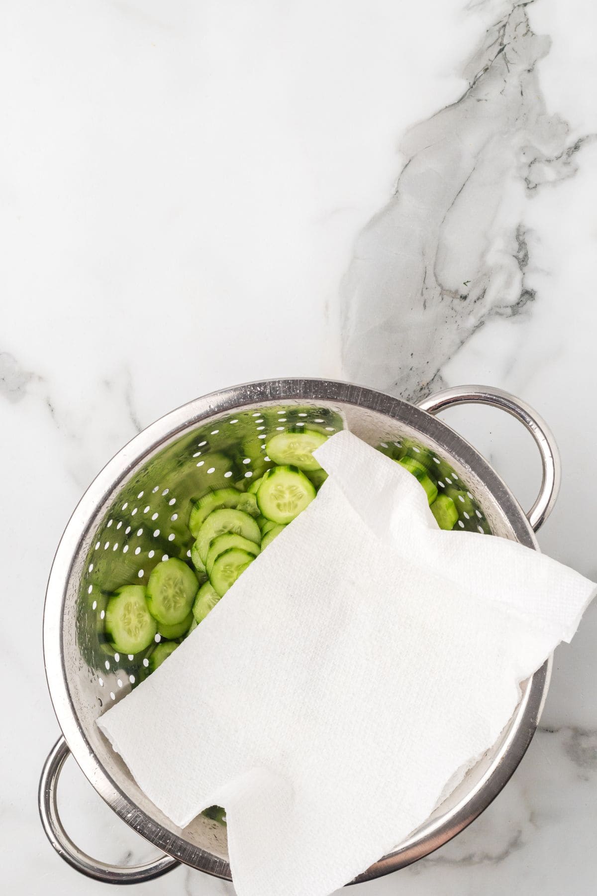 A metal colander containing sliced cucumbers partially covered with a white paper towel, placed on a white marble surface.