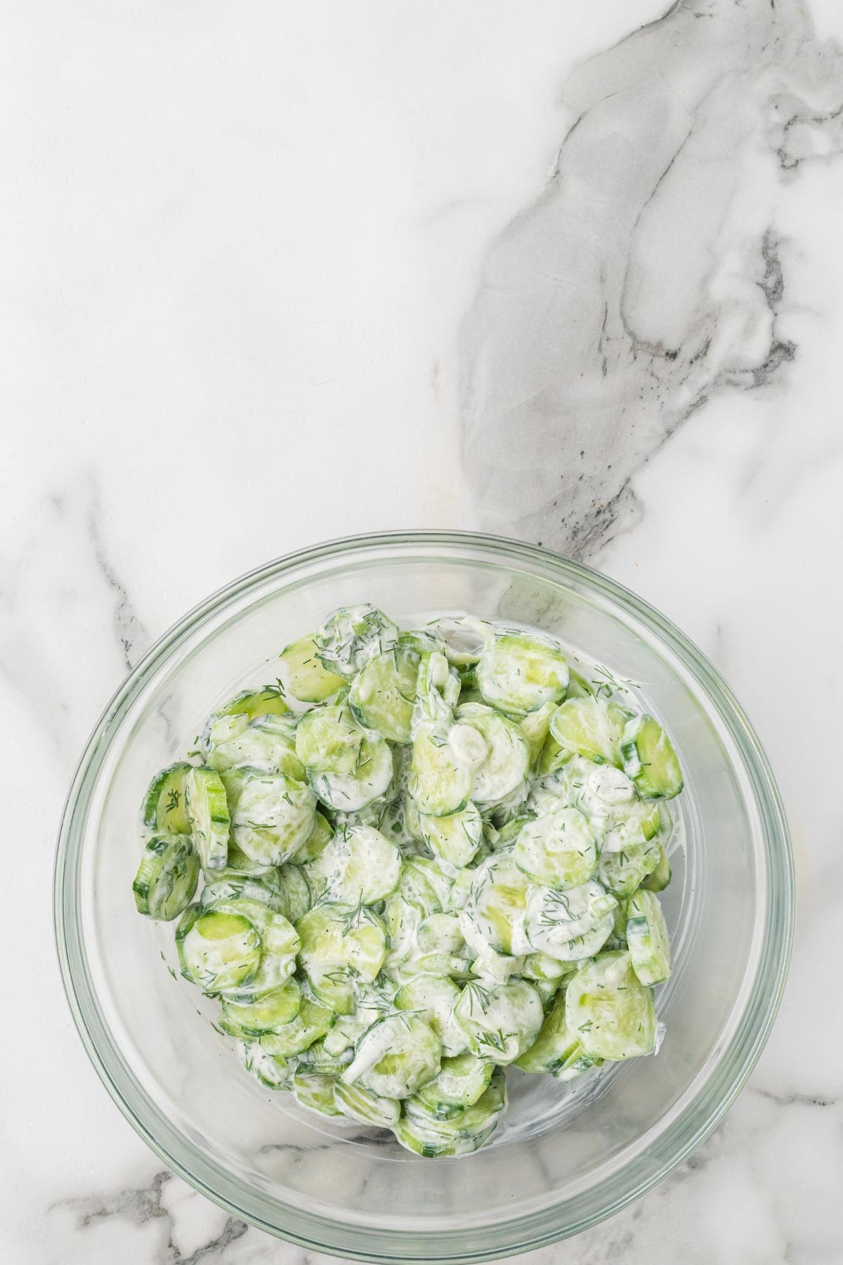 A clear glass bowl filled with sliced cucumbers coated in a creamy white dressing, sitting on a white marble surface.