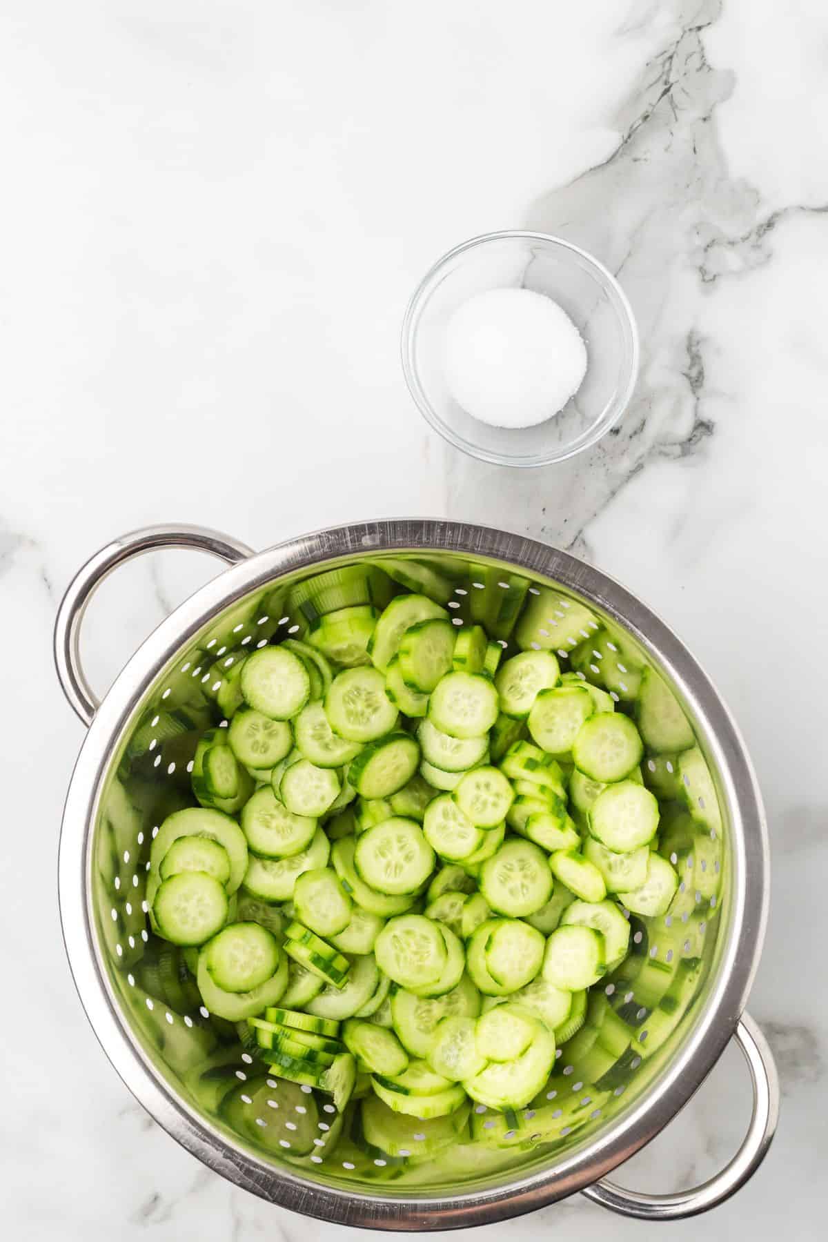 A metal colander filled with sliced cucumbers sits on a white marble surface, with a small glass bowl of white granulated salt placed above it.