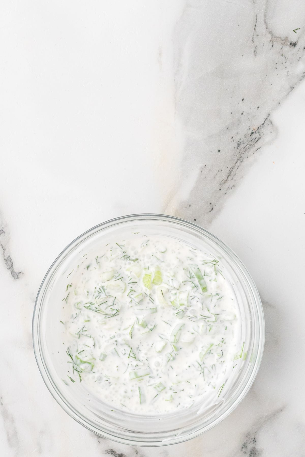 A clear glass bowl filled with a creamy white sauce containing visible green herbs and small vegetable pieces, placed on a white and gray marble surface.