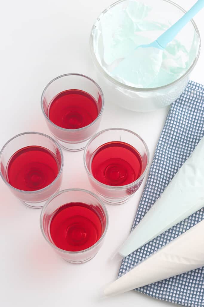 Four glasses of red gelatin, a bowl of whipped topping, and two piping bags on a blue checkered cloth.