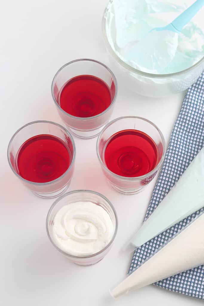 Four glasses of red gelatin, one topped with whipped cream, with piping bags and a bowl of whipped cream nearby.