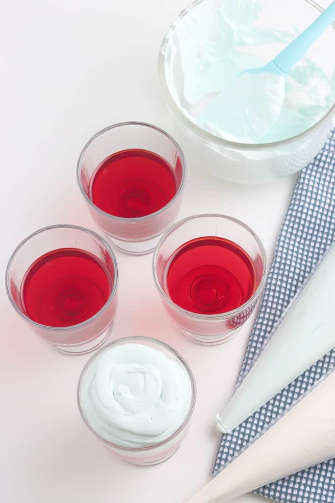 Four glasses of red gelatin, one topped with whipped cream, with a bowl and piping bags nearby on a checkered cloth.