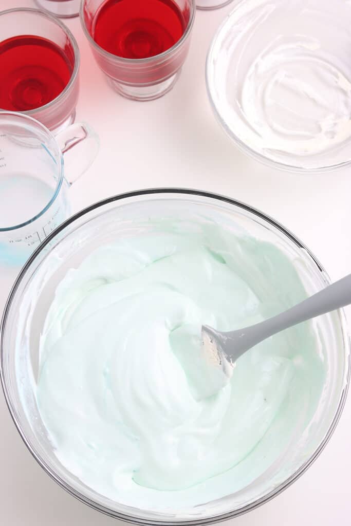 A bowl of fluffy, pale blue whipped mixture with a spatula, near cups of red gelatin and empty bowls.